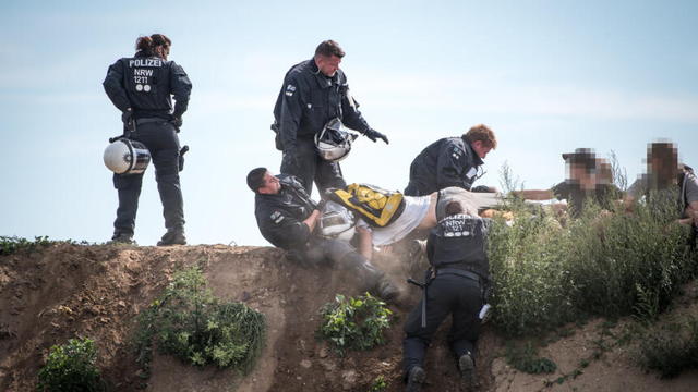 Mehrere Polizist*innen auf dem Hang. zwei stehen, einer fasst liegend am Boden an einem auf dem Bauch liegenden Menschen zerrend, hinter dem Menschen sitzen drei weitere (Gesichter unkenntlich). Sieht aus als versuchen sie den Menschen festzuhalten. Ein weiterer Polizist kniet gebückt am Boden. Eine Polizistin von hinten schaut mit Armen in der Hüfte zu der Sitation.