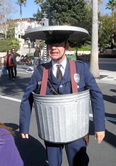 Man dressed up as a policeman wearing part of a trash can around him.