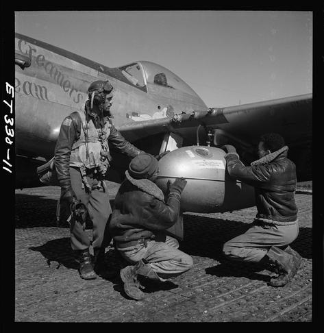 A black and white photo of three men working on a fighter jet, with the text "Creamers Dream" visible. One man is wearing an aviator helmet while another holds a fuel canister attached to the aircraft's wing. The third person appears to be assisting in securing or adjusting something related to the tank attachment.
The caption indicates that this image depicts Tuskegee airman Edward C. Gleed, along with two unidentified crewmen working on a P-51/D Mustang named "Creamer's Dream" at Ramitelli, Italy during March 1945. The photograph is part of an exhibit or collection related to the 332nd Fighter Group pilots and was provided by Tony Frissell from Images.LOENER.nl with reference number Gled-678a_07.