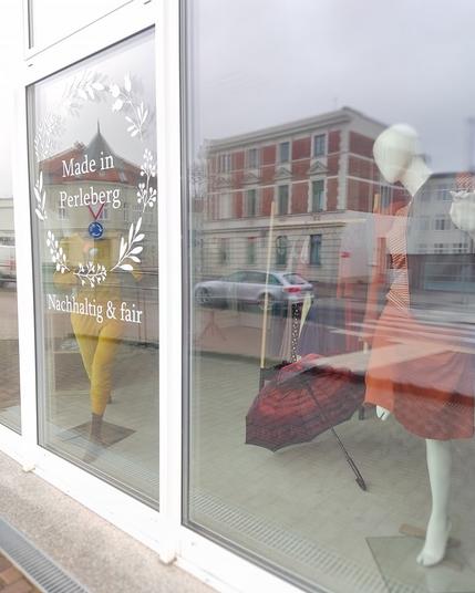 Blick ins Schaufenster von Frija Omina. Hinter den Glasscheiben stehen zwei Schaufensterpuppen. einmal mit rostfrbenen Rock und dazu farblich passender Weste und einmal in einem gelben Nicki-Anzug. Am Fenster steht Schrift "Made in Perleberg, nachhaltig & fair". In der Scheibe spiegelt sich die Straße mit einem Klinkerbau auf der gegenüberliegenden Straßenseite und einem silbernen Auto.