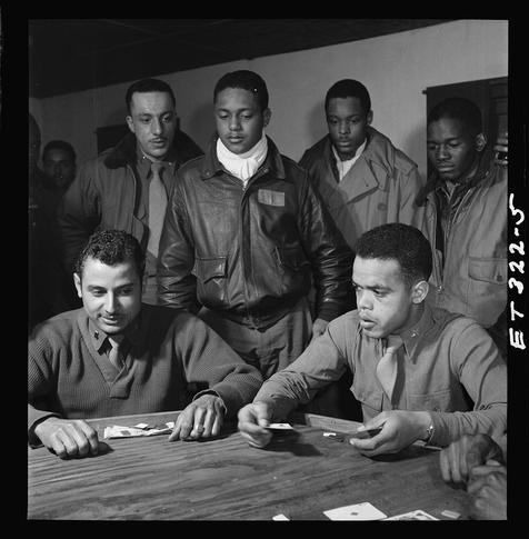 The black and white photograph captures a group of African American men, likely members of the Tuskegee Airmen. They are gathered around a table in what appears to be an officers' club during evening hours. The primary focus is on four seated men who seem engrossed in playing cards. One man sits prominently at the forefront with his hands poised over the deck as if he's about to play or has just played.

Behind him, three more men are observing closely; one of them appears particularly interested, possibly contemplating a move for himself. All individuals exhibit serious expressions and concentrated demeanors suggesting they're deeply engaged in their card game.
In addition to those seated around the table, there is another man standing behind the group on the right side with an attentive stance as he watches the proceedings at the table.

The men are dressed casually yet uniformly; most wear a mix of jackets or blazers over long-sleeved shirts paired with ties. Their attire suggests they're in military uniform style but not necessarily combat-ready gear, indicating that this might be a social gathering rather than a formal operational setting.
In summary, the photograph encapsulates an intimate moment among members of the Tuskegee Airmen as they partake in leisurely activities such as playing cards within their community space.
