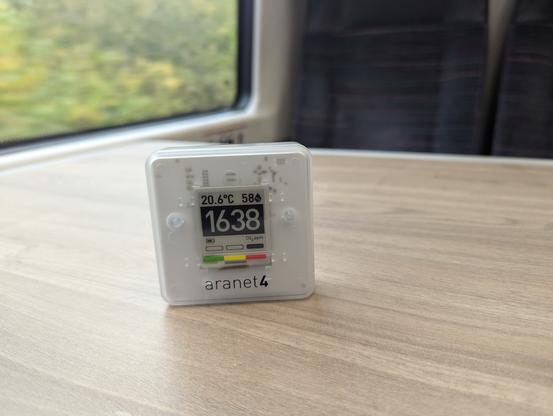 Aranet4 CO₂ monitor sitting on a table in a train. Greenery is visible out of the train window. The CO₂ level is showing as 1,638 PPM.