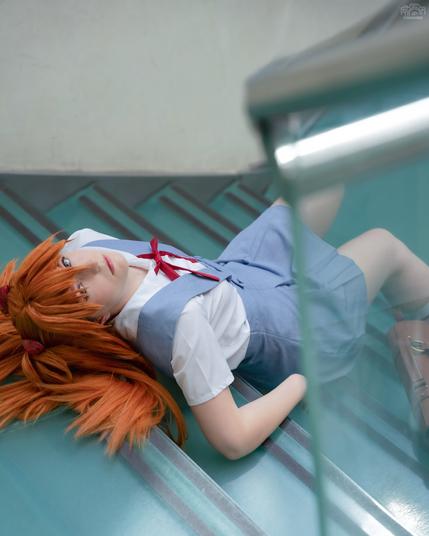 A cosplayer dressed as Asuka Langley from Neon Genesis Evangelion lies on the floor of a modern staircase with a glass railing. She wears Asuka's iconic school uniform with a white shirt, blue jumper, and red bow, with her long red hair fanned out behind her. Her expression is calm, staring upward, while the angle of the photo adds a dynamic, tilted perspective.