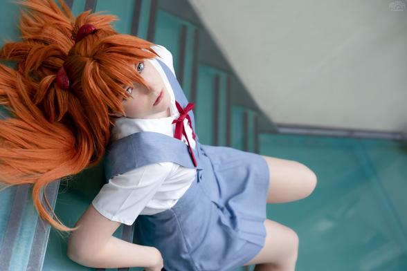 A cosplayer dressed as Asuka Langley from Neon Genesis Evangelion is lying on a staircase, leaning slightly upwards while gazing directly at the camera with a focused expression. She wears Asuka's recognizable school uniform with a white blouse, blue jumper, and red ribbon, her vibrant red hair styled in twin tails fanned out dramatically. The tilted angle of the photo and glass surface of the stairs add a dynamic touch to the scene.