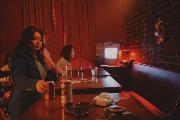 Picture of several people in a bar in a booth. One woman is looking down and reaching for a beer in the foreground. Another in the background is playing Mario kart on a small CRT television. The light from the television is illuminating her face.