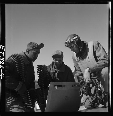 The image is a black and white photograph showing three men in what appears to be military attire, possibly pilots. They are gathered around an open briefcase or box with various objects inside that could potentially be ammunition belts. The man on the left wears heavy winter gear including a fur-lined coat and carries additional rounds of ammunition strapped across his chest. He is wearing goggles and has a cap pulled down over his eyes.
The middle man, also in cold weather attire, seems to be closely examining something within the box while interacting with one of the other men. The third man on the right is seated and appears more focused on the contents inside the case as well, holding what looks like an oxygen mask connected by a hose which suggests he may have been flying or preparing for flight.
All three individuals are wearing caps that indicate their affiliation to military units. Behind them, there's a vast expanse of clear sky with no visible landmarks, hinting at this scene being outdoors in open space possibly near the runway during daytime.