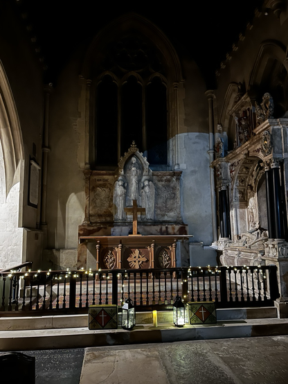 The altar view of an old church, heavily decorated with an effigy of a reclining man on the right. He is wearing a ruff and expensive clothes, his tomb is richly gilded and opulent