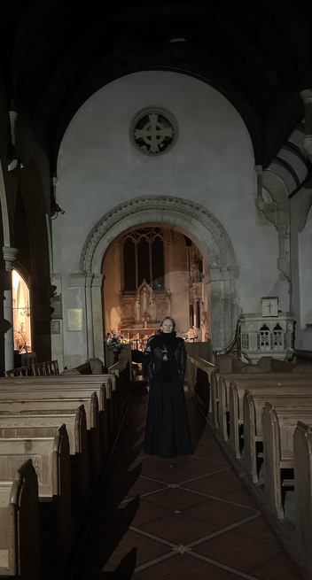 An indoor shot of a church altar, with a rounded doorway. In front stands a person slightly smiling wearing all black long skirt and a big scarf. You can see rows of pews on both sides