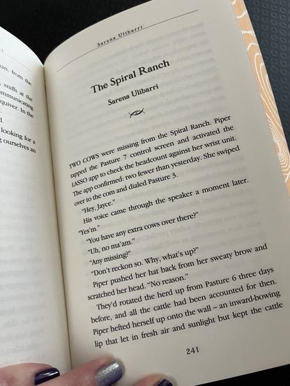 Picture of a book page held open. Text: The Spiral Ranch, Sarena Ulibarri. Two cows were missing from the Spiral Ranch. Piper tapped the Pasture 7 control screen and activated the LASSO app to check the headcount against her wrist unit. The app confirmed: two fewer than yesterday. She swiped over to the com and dialed Pasture 3.  
“Hey, Jayce.”
His voice came through the speaker a moment later. “Yes’m.” 
“You have any extra cows over there?”
“Uh, no ma’am.”
“Any missing?”
“Don’t reckon so. Why, what’s up?”
Piper pushed her hat back from her sweaty brow and scratched her head. “No reason.”
They’d rotated the herd up from Pasture 6 three days before, and all the cattle had been accounted for then. Piper hefted herself up onto the wall—an inward-bowing lip that let in fresh air and sunlight but kept the cattle