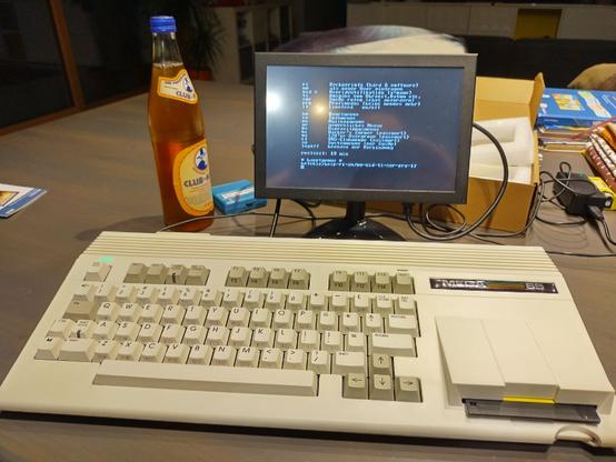 A Mega65 Computer on a Table, connected to a small display and a bottle of Mate next to it. The display shows white text on black background, apparently a connection to an old style Bulletin Board System