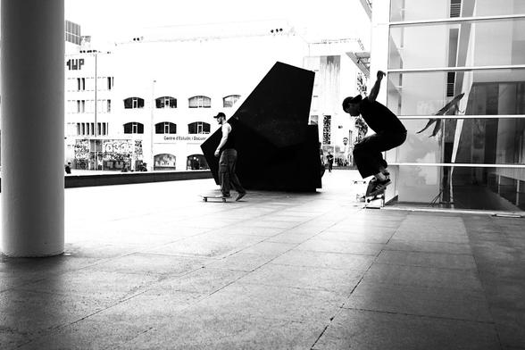 Black and white picture of a skateboarder performing an Ollie on a smooth stone surface. In the background a black stone sculpture can be seen, to the left a white pillar, to the right a glass facade