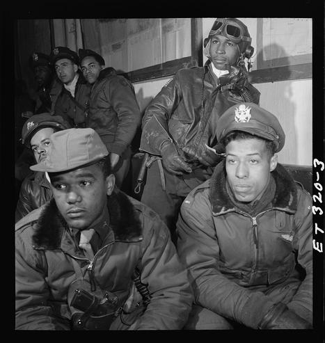 The image is a black and white photograph depicting several African American men dressed in military attire, most likely the Tuskegee Airmen. They are seated closely together with focused expressions on their faces as if they're listening to someone or waiting for an order. The front row consists of four men wearing bomber jackets with fur collars; two have a badge indicating rank and insignia, suggesting that one is a pilot while another could be part of the ground crew or support staff. In the background are more individuals in similar attire but without badges visible.

The setting appears to be an indoor environment like a briefing room within Ramitelli airbase during March 1945 as per historical records and caption information provided alongside this image, which is sourced from the Tuskegee Airmen's archive. The men’s postures and expressions convey seriousness and attentiveness toward their duties or mission briefings.

This photograph captures an important piece of history regarding African American pilots during World War II who broke racial barriers in aviation.