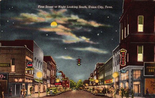 A night scene in a small town. You are looking right down the street. Lights, cars, and people are all around. There are stars, and a full moon dotted with clouds.