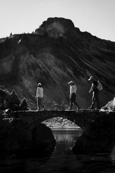 Three people walking across a stone archway over a creek with a lake and mountain in the background.