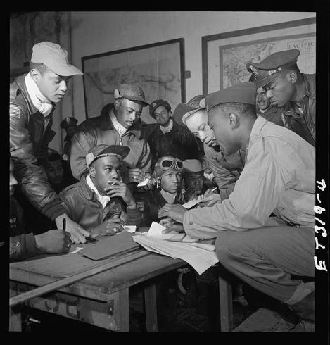 The image depicts a group of African American pilots, known as the Tuskegee Airmen, gathered around a table in an indoor setting. They are dressed in military attire and some wear aviator goggles or caps typical for their era. The men appear engaged with paperwork on the table, possibly discussing operations or flight plans.

The room is adorned with maps of various regions including "THE PACIFIC AT EAST" hanging on the wall behind them, indicating that they may be strategizing future missions in World War II. Their focused expressions and collaboration suggest a serious atmosphere as they prepare for their duties during this time period.

Notable details include an individual labeled Jimmie D. Wheeler wearing goggles with his name written below him along with "San Francisco, CA, Class 44-B". Another is Joseph L. Chineworth from Memphis, TN, Class 44-E; and Woodrow W. Crockett also identified as being at Ramitelli, Italy in March of that year.

The photograph captures a moment highlighting the Tuskegee Airmen's determination and contribution to WWII while showcasing their camaraderie and teamwork amidst challenging circumstances.