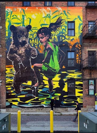 Streetartwall. The beautiful mural of a little girl with a bear was sprayed/painted on a red brick wall with three windows of a multi-storey house. The background is a marshland landscape with plants and water painted in bright yellow, green and orange. The little girl with long brown hair is wearing a short green dress and jumping around in the glittering water. Her ponytail is blowing and she casts a furtive glance over her shoulder. A little brown bear stands next to her and dances with her. A wonderful energy emanates from this positive mural.
Info: Pipsqueak was here!!! is an Amsterdam-based artist duo driven by a strong passion for all life on planet Earth. Animals are not metaphorical placeholders for humans, they are animals. The children are children.