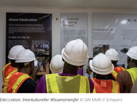 Foto einer Gruppe Menschen, von hinten gesehen, in Warnwesten und Schutzhelme gekleidet, die vor einer Reihe Informationstafeln zum Edo|Cation-Projekt stehen.