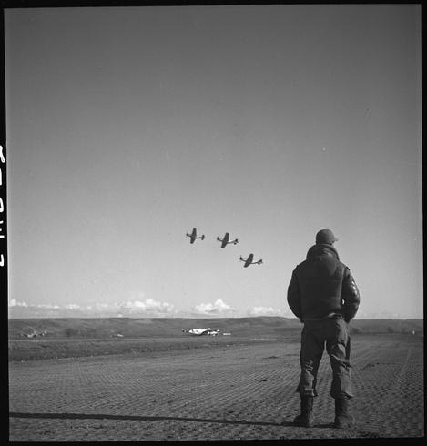A black and white photograph depicting an individual, likely a pilot or military personnel given the context provided. The person is standing on what appears to be a dirt field with sparse vegetation in the foreground. They are dressed in dark attire that includes a heavy jacket, trousers, boots, gloves, and headgear consistent with cold-weather gear.

Behind this figure stands an open expanse leading up to a clear horizon line where three aircraft can be seen flying in formation against a blue sky dotted with clouds. The planes resemble vintage military or fighter aircraft from mid-20th century design styles.

The overall mood of the image is contemplative and solitary, suggesting a moment reflective upon flight operations possibly during WWII given historical context provided by caption details pertaining to an unidentified Tuskegee airman in Italy March 1945.