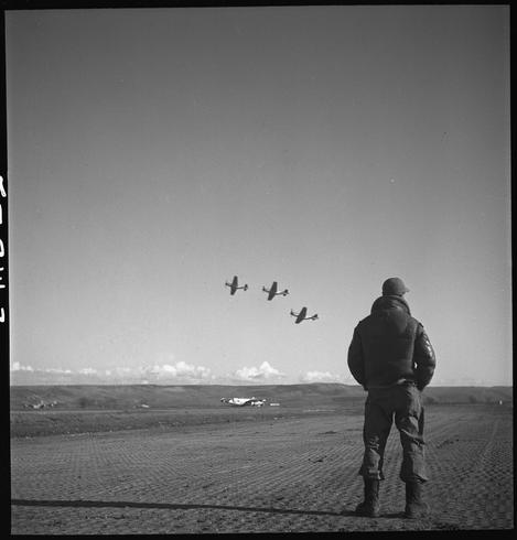 A black and white photograph depicting an individual, likely a pilot or military personnel given the context provided. The person is standing on what appears to be a dirt field with sparse vegetation in the foreground. They are dressed in dark attire that includes a heavy jacket, trousers, boots, gloves, and headgear consistent with cold-weather gear.

Behind this figure stands an open expanse leading up to a clear horizon line where three aircraft can be seen flying in formation against a blue sky dotted with clouds. The planes resemble vintage military or fighter aircraft from mid-20th century design styles.

The overall mood of the image is contemplative and solitary, suggesting a moment reflective upon flight operations possibly during WWII given historical context provided by caption details pertaining to an unidentified Tuskegee airman in Italy March 1945.