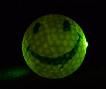 ein mit grinse Smiley Gesicht bemalter Golfball liegt auf dem Boden. In ihm steckt ein Knicklicht, das ihn hellgrün von innen beleuchtet