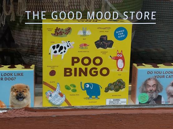 Lähikuva kaupan ikkunassa olevasta POO BINGO-pelistä. Keltaisen rasian kannessa on erilaisten eläinhahmojen piirroskuvia ja kakkakasoja. Kaupan ikkunassa teksti: THE GOOD MOOD STORE