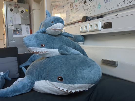 A big, a small and a blahaj stacked ontop of each other. In the background there are more sharks on the sofa or on the heater.