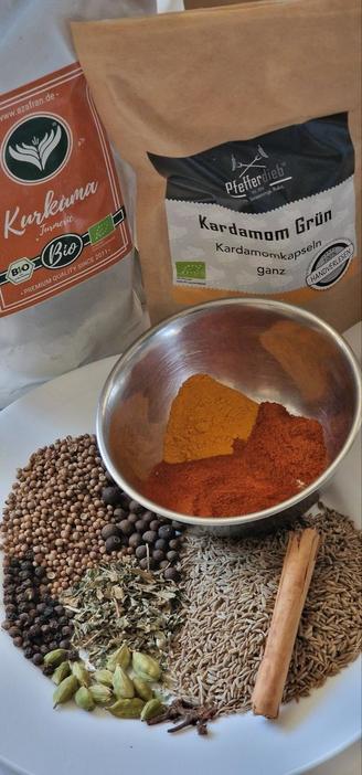 Mischung Berbere-Gewürz auf weissem Teller: schwarze Pfefferkörner, Piment, Zimtstange, grüne Kardamomkapseln, Koriandersaat, Kreuzkümmel, Bockshornkleeblätter, Nelken, edelsüßer Paprika, Chili, Kurkuma, Muskat.