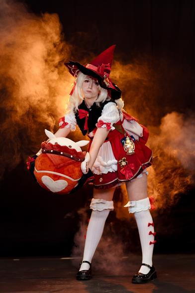 A cosplayer dressed as Klee from Genshin Impact is posing in an action stance, holding a large plush version of her iconic explosive "Jumpy Dumpty." The cosplay is highly detailed, featuring Klee's signature red and white adventurer's outfit with a pointy red hat adorned with bows and accessories. Smoke or fog surrounds the cosplayer, adding a dynamic and dramatic atmosphere to the scene, reminiscent of Klee's explosive abilities in the game.