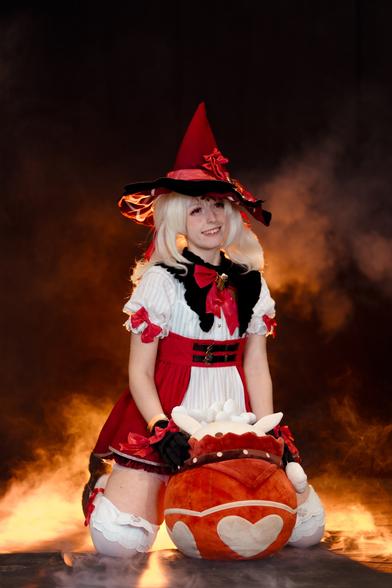 A cosplayer dressed as Klee from Genshin Impact is kneeling down with a big smile on her face, holding her iconic plush "Jumpy Dumpty" bomb in front of her. The outfit is Klee's vibrant red and white adventurer attire, complete with her large, pointy red witch hat adorned with ribbons and details. The background is filled with smoky effects, creating a playful yet fiery atmosphere, perfectly matching Klee's explosive personality.