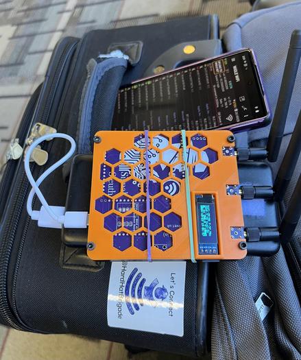An Android phone in a purple case running WigleWiFi and a SignalSleuth with an orange case are perched on a black suitcase with Hard Hat Brigade white and blue sticker stating “Let’s Connect”