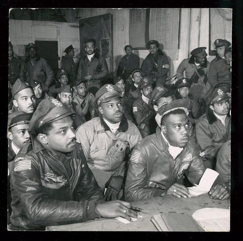 The image is a black and white photograph depicting several African American men in military uniforms, specifically those of the 332nd Fighter Group. They are seated at what appears to be a briefing room table or bench within an indoor setting that resembles a building with exposed brickwork on its walls.

Most individuals wear bomber jackets typical for pilots during World War II; many have patches and insignias indicative of their affiliation with this specific group, such as the American flag patch. Some men are wearing hats bearing distinctive emblems which might signify rank or unit membership within the air force. The expressions range from focused to contemplative.

In addition to these seated figures, others stand in the background, suggesting a larger gathering beyond just those around the table. One can see items like notebooks on the tables and personal belongings among the group members.

The photograph carries historical significance as it likely documents one of many such briefings for fighter pilots during World War II era operations involving African American servicemen, known collectively as the Tuskegee Airmen due to their training in Tuskegee.