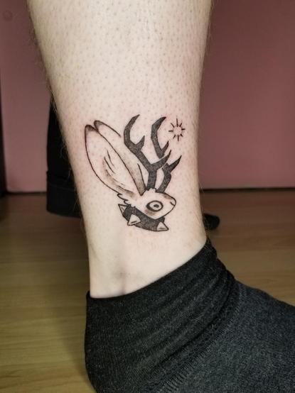 Photo of a tattoo I did of one of my "mini cryptids" flash designs. A lil jackalope head, with a large spiked collar on, and a soft little star/sparkle right in front of their antlers.