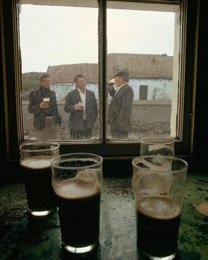 Photograph. An old color photo of three men drinking Guinness in front of a pub in Ireland. The photo is taken looking out of the dark pub towards the window. There are four half-full pint glasses on the green windowsill (or table) in the foreground, but the view is through a two-paned, somewhat dirty wooden window to the outside. Three men, all dressed differently in green and gray tweed (with white shirts or black sweaters and caps), stand in front of the window, chatting and comfortably drinking a glass of Guinness. A flat, rural estate can be seen behind them. Such a charming old photo with a lot of flair from the "good old days", if you can call three men drinking "charming".
Info: The photo comes from an article about Ireland in the magazine "Nationale Geographic" from 1971.