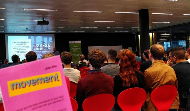 People sitting in red chairs listening to a presentation at the start of a Hackathon. In the foreground a pink leaflet with the word "movement" on it.