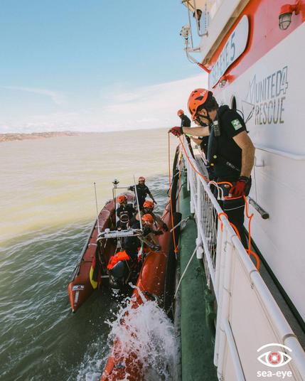 Ein Mensch der sea-Eye Crew an der Reling des Schiffes, welcher gerade ein Rettungsboot ins Meer lassen in dem Boot sitzen ebenfalls vier Menschen der Crew.