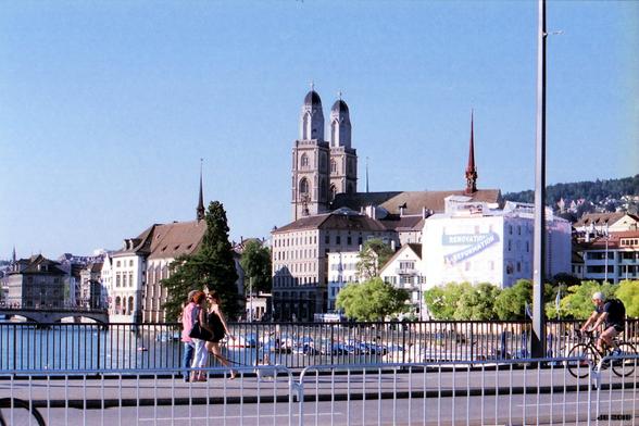 Farbfoto, aufgenommen von einer Brücke über die Limmat auf einen Teil des Stadtzentrums von Zürich, Schweiz. In Bildmitte die Türme des romanischen Grossmünsters. Im Vordergrund Passanten und ein Fahrradfahrer auf der Brücke. Leica R3 mit 35 mm Summicron auf Fuji PRO 400-Film.