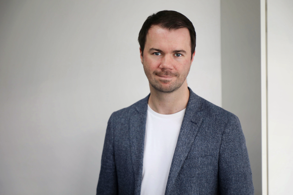Porträtaufnahme von Marc Philipp Janson. Er steht vor einer weißen Wand. Zu sehen sind Kopf und Oberkörper. Er hat dunkle kurze Jahre und einen Drei-Tage-Bart, trägt ein grau-meliertes Sakko, darunter ein weißes T-Shirt. Foto: Pädagogische Hochschule Karlsruhe