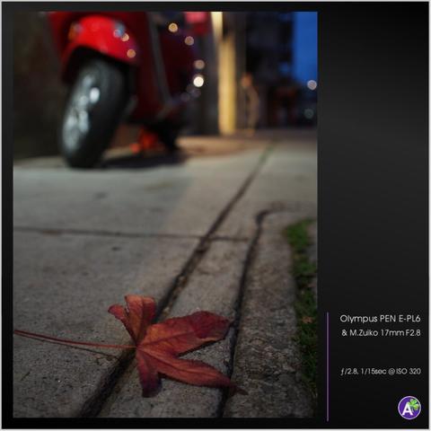 a color outdoor photograph. a red maple leaf sits on a city sidewalk. behind it, defocused slightly in the background, sits a red Vespa-type scooter. the scooter and leaf are the same shade of red. streetlights and specular highlights behind the scooter are defocused so that they appear only as small circles of light and color. there are no animals or people in this photograph