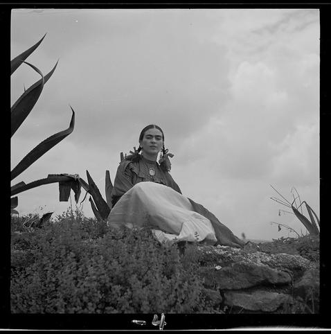a black and white photo of a woman in native american style dress, with large earrings and long dark hair is seated on rocks near several tall plants. she gives the "peace" sign to the camera as her expression looks off into space towards something beyond what we can see or imagine
