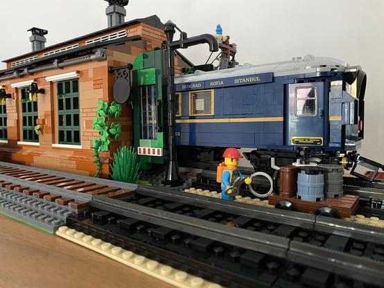 Ein blauer Salonwagen aus Lego ragt halb aus einem altertümlich-kleinen Lokschuppen heraus.