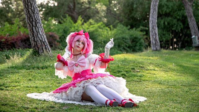 A cosplayer dressed as Madoka Kaname from Puella Magi Madoka Magica sits elegantly on a grassy field, surrounded by trees and greenery. The outfit is a highly detailed, frilly pink and white dress with large bows and puffy sleeves, mimicking Madoka's magical girl attire. The cosplayer has pink hair styled in twin pigtails, matching the character's signature look. With delicate gloves and expressive body language, they capture Madoka's innocence and charm. The bright sunlight filtering through the trees enhances the serene, fairy tale-like atmosphere of the scene.