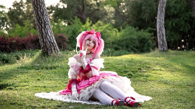 A cosplayer dressed as Madoka Kaname from Puella Magi Madoka Magica poses gracefully on a grassy field. Wearing a detailed pink and white frilly dress with bows, lace, and ribbons, they capture the innocence and magical girl essence of the character. The pink twin pigtails, white gloves, and vibrant accessories complete the look. Sitting on the ground with a hand gently raised to their face, the cosplayer conveys a serene and thoughtful expression. The natural sunlight filtering through the trees and the soft greenery around them adds to the whimsical, dreamlike atmosphere of the scene.