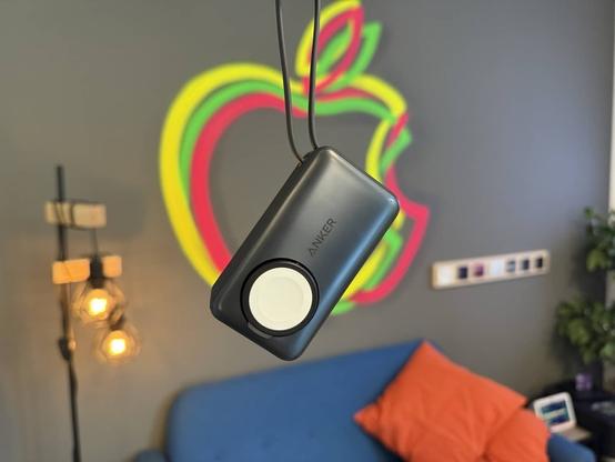 Anker MagGo Powerbank für Apple Watch baumelt an ihrem USB-C-Kabel. Im Hintergrund ein dreifarbiges Apple-Logo an der Wand sowie ein blaues Sofa darunter.