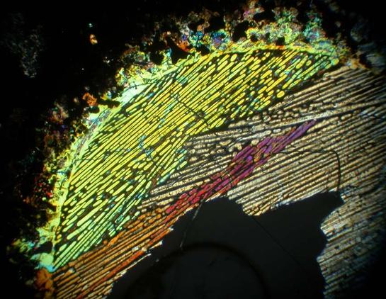 A spherical chondrule (solidified melt formed at the start of our Solar System) in cross polarized transmitted light. Thin blades of quickly crystallized minerals give color. Horizontal field of view approximately 0.7 mm.