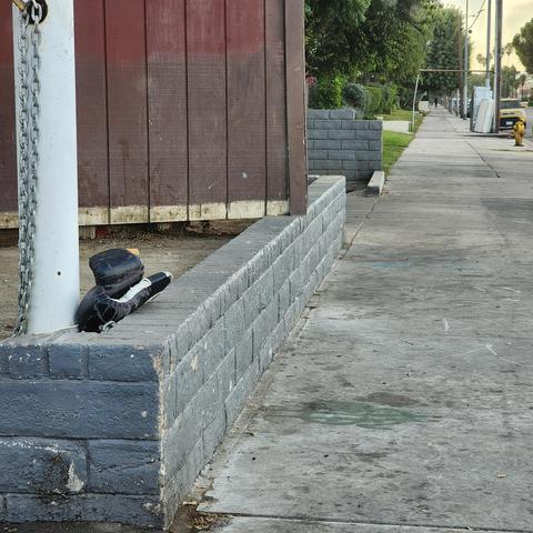 A sidewalk along sepulveda blvd in the early morning, a short wall runs along the sidewalk painted dark grey, a white pole at the corner of the wall and driveway near the camera, and an inexplicable pair of heavily used black boxing gloves lain atop the wall against the pole. Nobody else around.