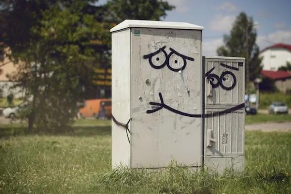 Two grey utilities boxes stand side by side in a green field. One has sad eyes graffitied on it. The other has warm caring eyes, and arms hugging the first one,  drawn around both sides.