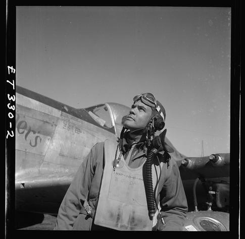 The image is a black and white photograph showing an individual, presumably Colonel Benjamin O. Davis, standing next to what appears to be an airplane from World War II era. The person is wearing flight gear including goggles on the head, earphones with microphone attached around his neck, pilot's jacket over casual clothing, and a protective apron labeled "NSP-10-G-B" along with some other illegible text.

He has dark hair under the helmet that’s not covered by it. His gaze is directed upwards towards something outside of frame which seems to capture him in mid thought or anticipation. The aircraft behind him bears visible markings and numbers, but they are partially obscured; however "738012" can be discerned.

The photograph appears old with speckles indicative of age on the film negative. It's taken outdoors during daylight as indicated by the clear sky above which is devoid of any clouds or birds, suggesting a dry atmosphere typical for flying conditions.