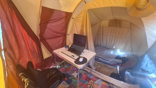 A fisheye lens shot of the inside of the tent. In the foreground we see a desk with computer on a colorful carpet that was draped over the cart before. In the background, in the inner tent, a red sleeping mat with a large gray sleeping bag and some clothes scattered about.