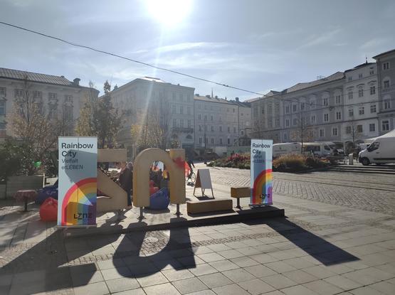 The main square of Linz on a sunny day. There is a foto spot where people can be the "i" in "Linz". Before the "L" and the "Z" there are rainbow banners saying "Rainbow city". Behind an information stand is being set up as an opportunity to interact with the public.