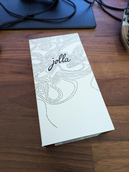 A picture of a white box with 'Jolla' written on it in front of a thin line pattern.  It's on a dark brown desk.